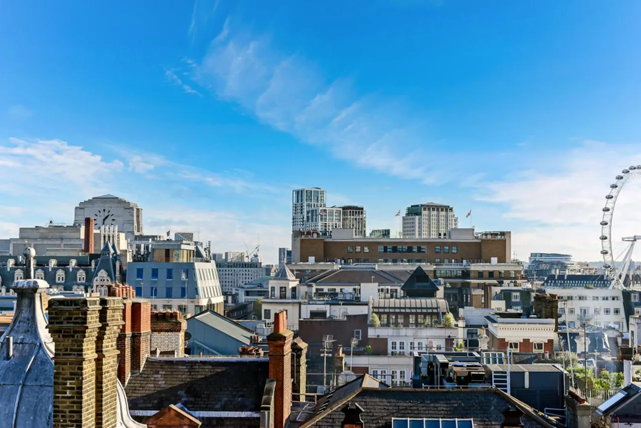 View over rooftops, chimneys and city buildings toward the London Eye under a blue sky.