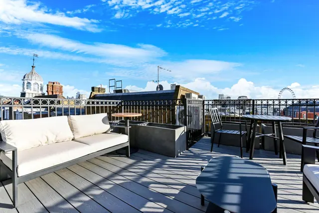 A rooftop terrace with a white sofa, a low table, and dark metal dining chairs, with the London Eye and city buildings visible beyond the railing.
