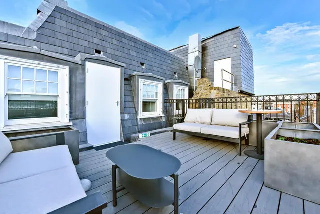 A roof terrace with grey decking, a white sofa, grey tables and planters, with a railing and rooftops beyond under a blue sky.