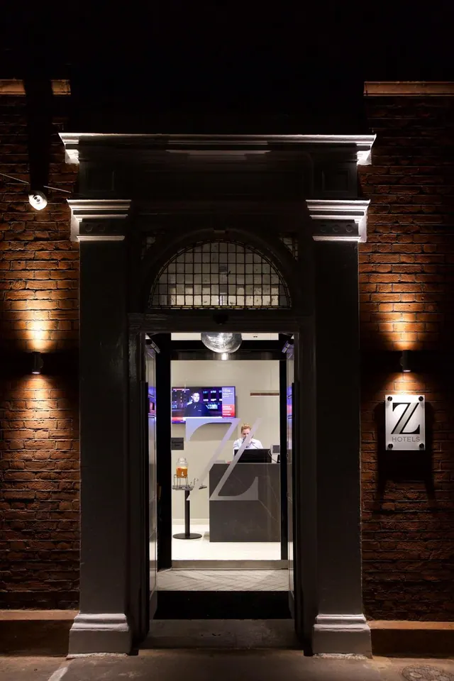 Night view of a brick building entrance with a dark framed doorway opening onto a reception area, where a staff member sits behind a desk beneath a wall-mounted TV.
