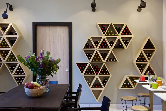 A dining area with geometric wooden wine racks mounted on the wall, a dark wooden table with a fruit bowl and a flower vase, and stools and chairs.