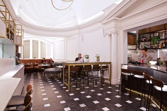A bar and seating area with a domed white ceiling, black and white tiled floor, high tables, and a bartender working behind the counter.
