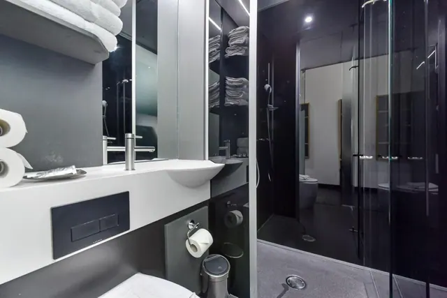 A bathroom with a wall-mounted basin, a dark flush plate, folded towels on open shelving, and a glass-enclosed walk-in shower.