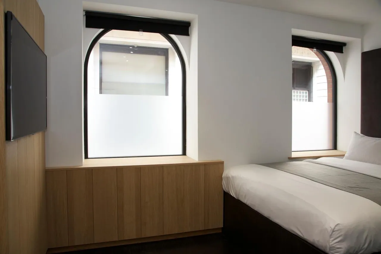 A bedroom with a double bed, a wall-mounted television, and arched windows with frosted lower panes above a wooden cabinet.