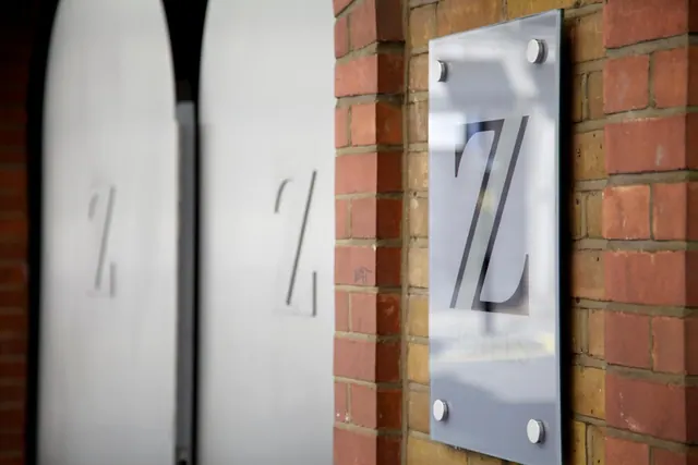 A frosted glass sign with a large letter Z is mounted on a brick wall beside arched white entrance doors.