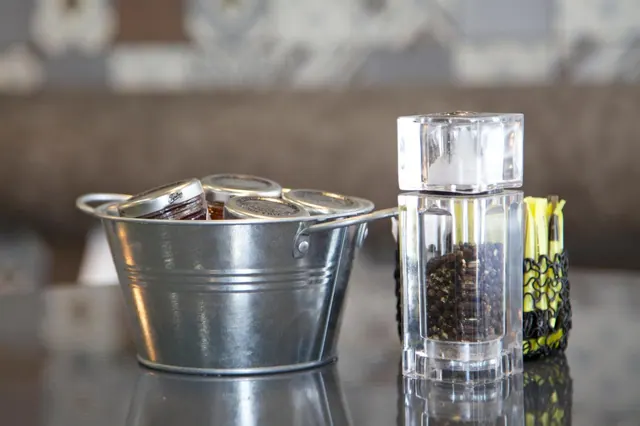 A galvanized metal bucket of jam jars and an acrylic salt and pepper grinder stand on a dark table beside a wire holder of sugar sachets.