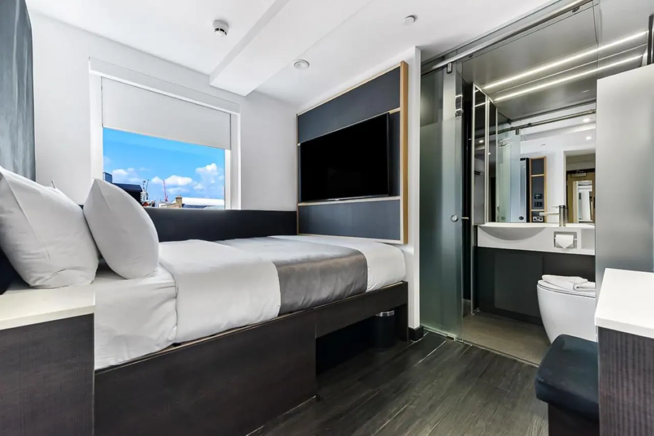 A hotel bedroom with a double bed, a wall-mounted television and a window showing blue sky and rooftops, with an adjoining bathroom visible on the right behind a glass sliding door.