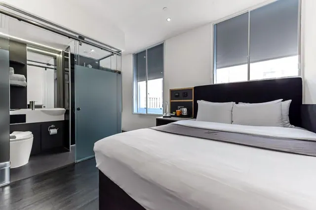 A bedroom with a king bed, with a glass-walled shower, sink and toilet visible in the adjoining bathroom at left and a kettle on the bedside table.