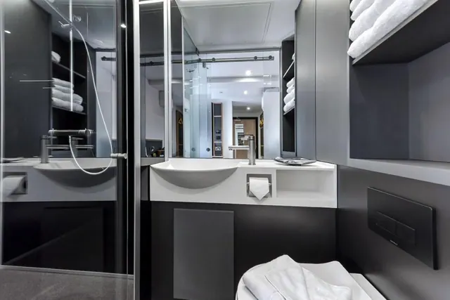 A black and white bathroom with a glass-walled walk-in shower, a white wall-mounted sink with a towel roll, and folded towels on open shelves.