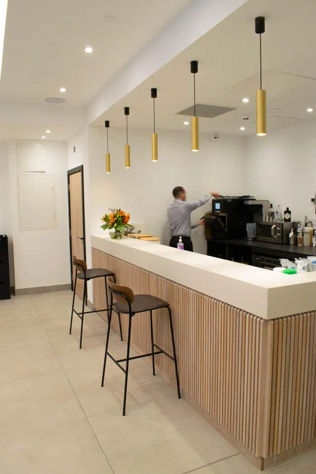 A staff member works behind a timber-fronted counter beside a coffee machine, with bar stools in front and gold pendant lights hanging above.