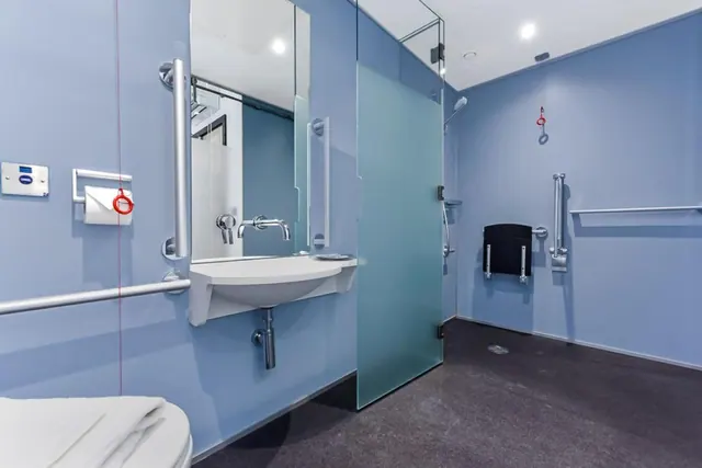 An accessible bathroom with blue walls, a wall-mounted sink with grab rails, a frosted glass walk-in shower and a fold-down shower seat, with a toilet partly visible in the foreground.