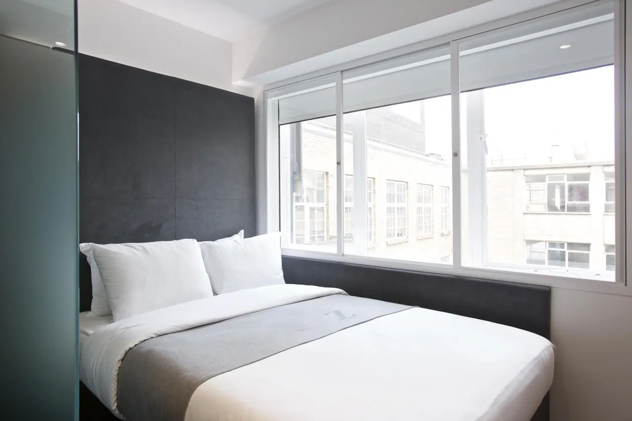 A double bed with white linen and a dark upholstered headboard sits beside a large window looking onto neighbouring brick buildings.