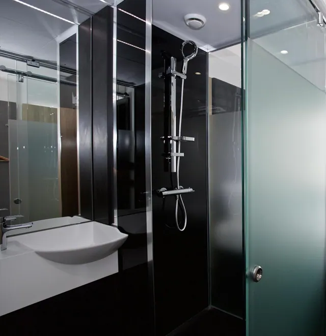A black-walled bathroom with a white wall-mounted basin, a handheld shower on a slide bar, and a frosted glass shower enclosure.