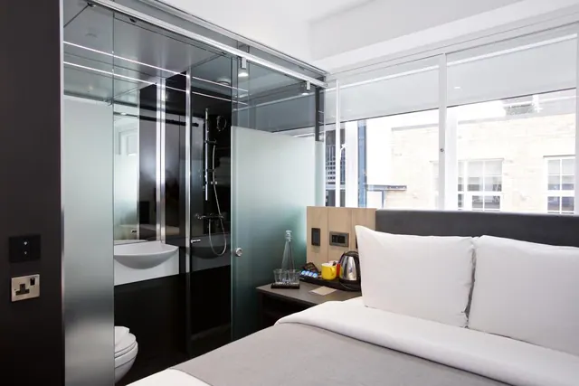 A bedroom with a double bed beside a glass-walled bathroom containing a walk-in shower, sink and toilet, with a window overlooking a brick building.