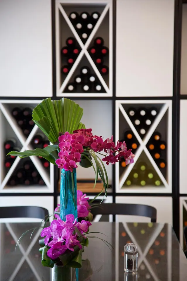 A dining table holds a blue vase of pink orchids and palm fronds, with chairs and a wall of wine bottles in racks behind it.