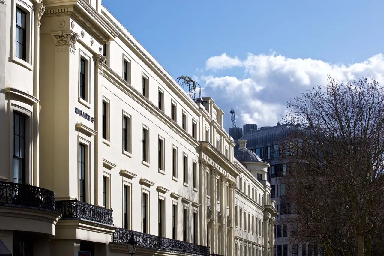 Exterior of a cream stucco period building with iron balconies and a bare tree on the right, under a partly cloudy blue sky.