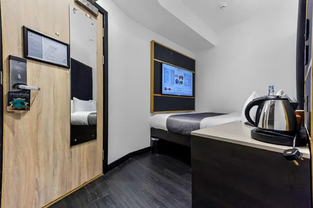 A small hotel bedroom with a single bed, a wall-mounted television, a kettle on a dresser, and a wooden door with a keycard lock and mirror.