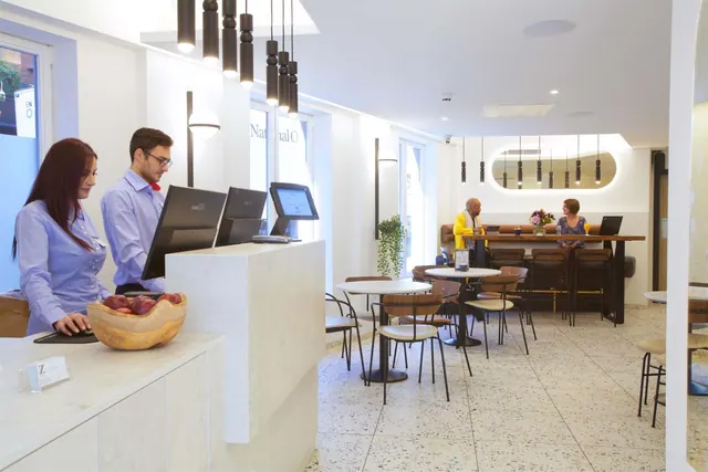 Two staff stand behind a white reception desk with monitors and a fruit bowl, beside a café seating area where a few guests sit.