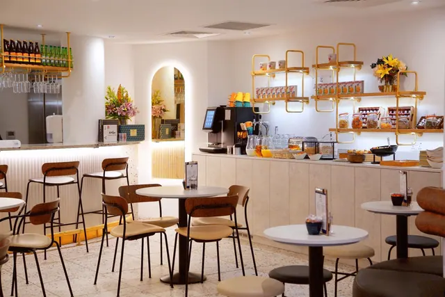 A breakfast area with round tables and chairs, a coffee machine, juice jugs and cereal dispensers on a counter under gold shelving, with a stainless steel fridge at left.