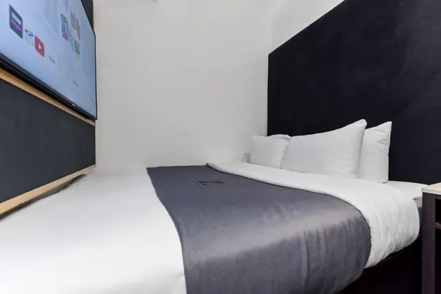 A double bed with white linen, a grey throw and a dark upholstered headboard, with a wall-mounted television on the left.
