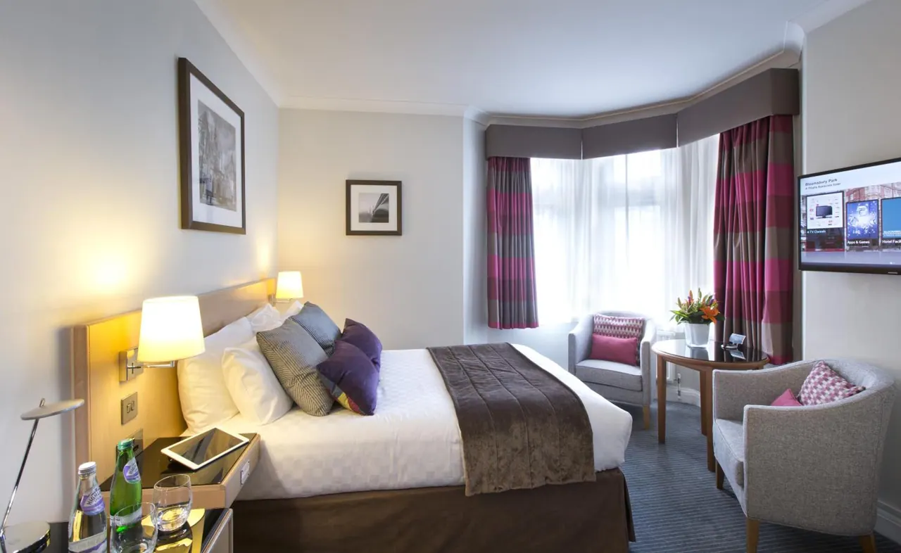 A double bed with purple cushions, bedside lamps, a bay window with striped curtains, two armchairs and a wall-mounted television in a hotel bedroom.