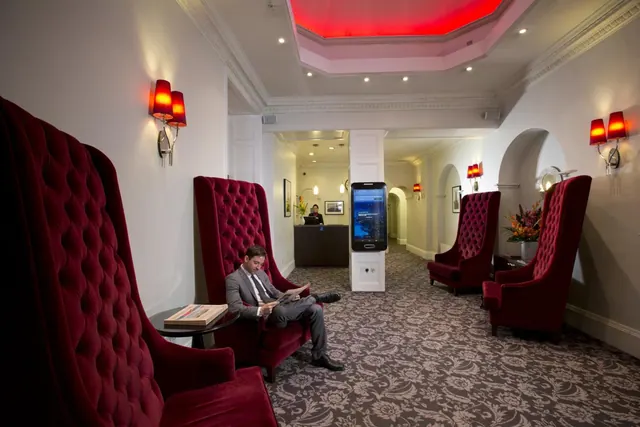 A man in a grey suit reads a newspaper in a tall red tufted armchair in a hotel lobby, with a reception desk and a touchscreen kiosk visible behind him.