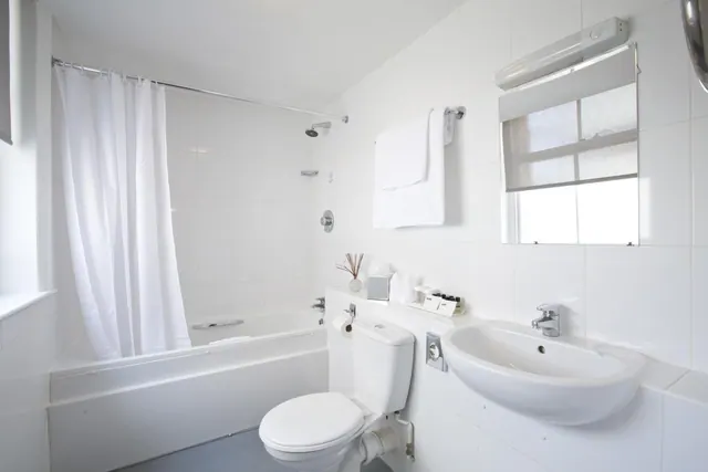 A white bathroom with a bathtub and shower over it behind a white curtain, a toilet, a wall-mounted sink, hanging towels, and a window with a blind.