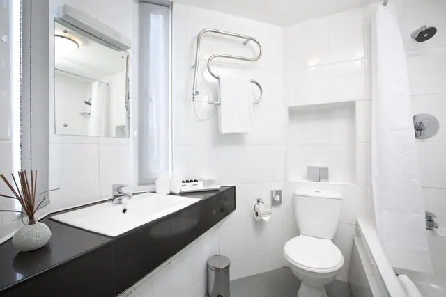 A white bathroom with a single sink on a black countertop, a toilet, a heated towel rail holding a towel, and a bath with a shower curtain.
