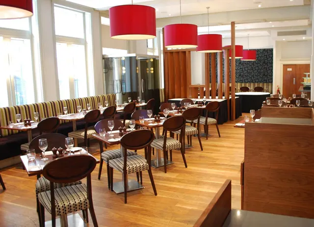 An empty restaurant dining room with tables set with wine glasses, red pendant lamps, patterned chairs and a striped banquette along tall windows.