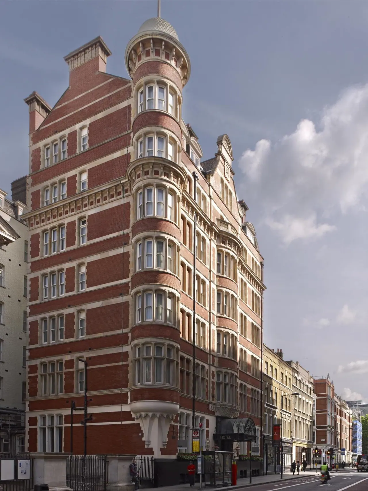Exterior view of a red brick Victorian building with a domed corner turret and bay windows, beside a street with pedestrians and traffic.