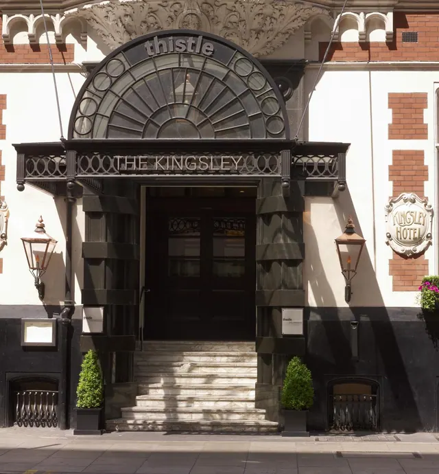 The front facade of a hotel building with an arched glazed fanlight above a dark entrance, marble steps, potted shrubs and a sign reading The Kingsley Hotel.