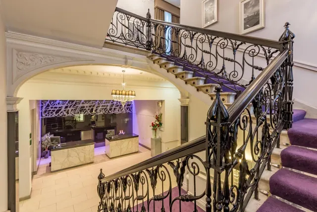 A staircase with an ornate wrought-iron railing and purple carpet rises beside an arched lobby with a reception desk and a chandelier.