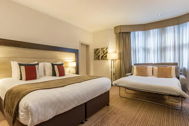 A bedroom with a large double bed with cushions, a pulled-out sofa bed beside a bay window with sheer curtains, a floor lamp and a striped carpet.