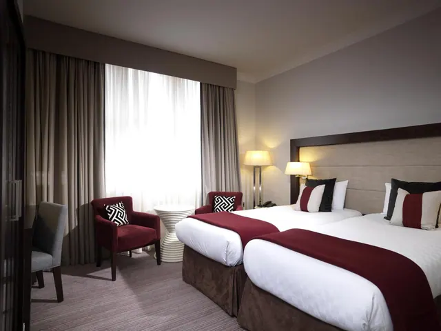 A hotel bedroom with two beds with red throws, red armchairs and a white side table beside a bright window with sheer curtains.