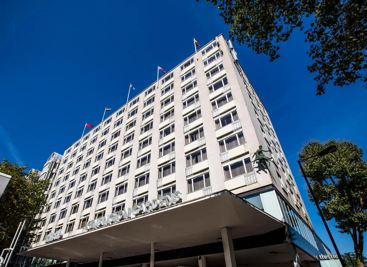 Low-angle view of a multi-storey hotel building with a cantilevered canopy bearing the Kensington Gardens sign, rooftop flags, and a clear blue sky.