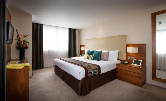 A king bed with a cream upholstered headboard and teal cushions sits between wooden bedside tables, with a wall-mounted television on the left and a tall mirror on the right.