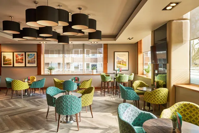 A lounge with patterned teal and green armchairs around small round tables, black pendant lights overhead, framed prints on the wall, a wall-mounted television, and a brick building visible through the windows.
