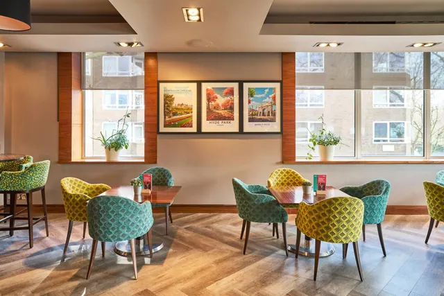 A seating area with patterned teal and yellow armchairs at wooden tables, framed London park posters on the wall, and windows looking out onto a brick building.