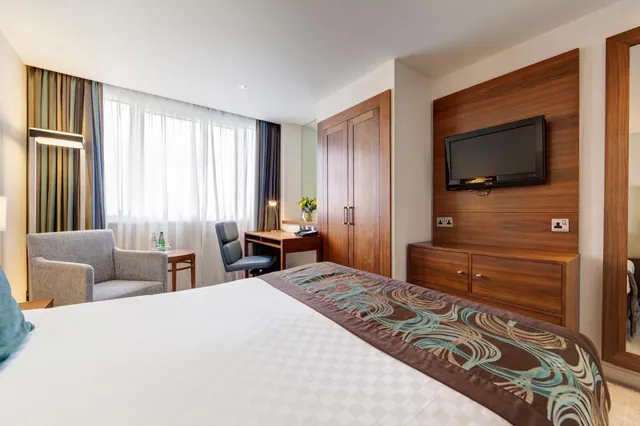 A hotel bedroom with a large bed, a wall-mounted television, a wooden wardrobe, a grey armchair and a desk by the window.