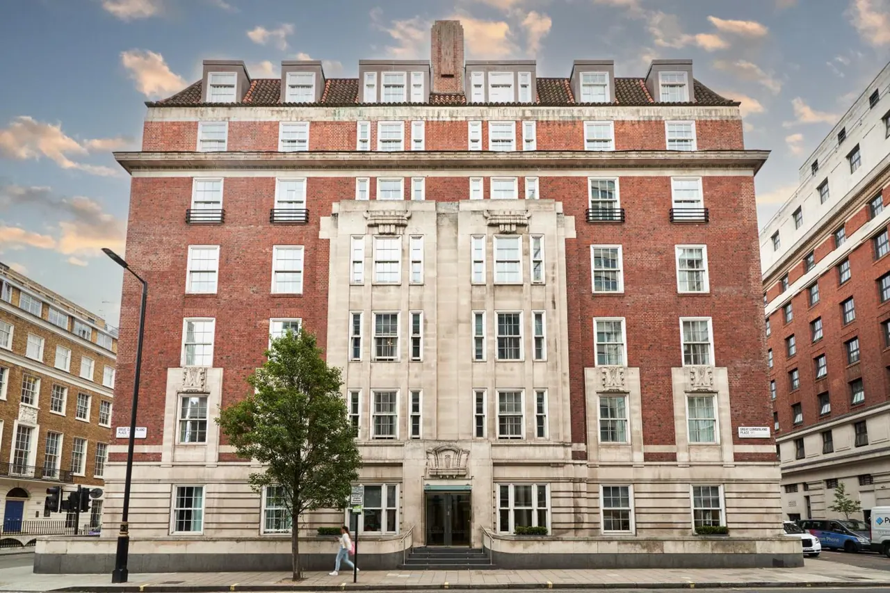 The front of a red brick building with a pale stone central section, seen from the street with a tree and a pedestrian in front.