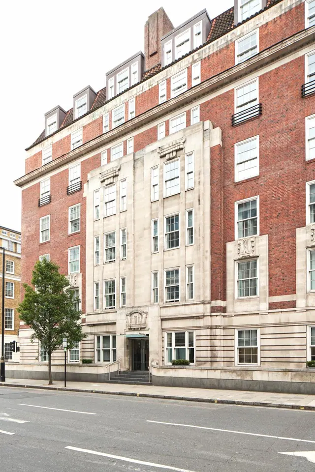 Street-level view of a red brick building with a pale stone central entrance bay, a canopy and steps up to the door, and a tree on the pavement.