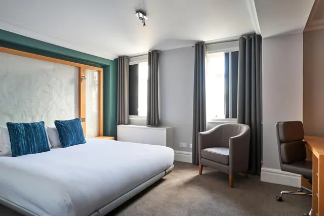 A bedroom with a made double bed with blue cushions, a grey armchair by the window, and a wooden desk with a chair.