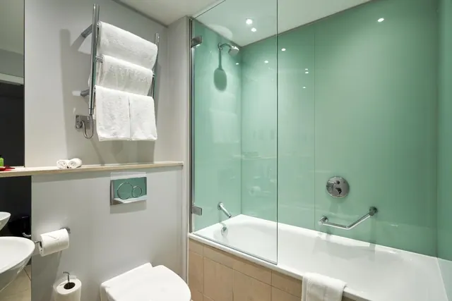 A bathroom with a bath under a glass screen, a shower head above it, a green glass wall, a heated towel rail, a grab rail and a toilet.