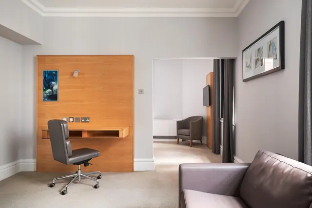 A hotel suite living area with a wood-panelled wall desk and leather office chair, a leather sofa in the foreground, and an armchair and wall-mounted television visible through an open doorway.