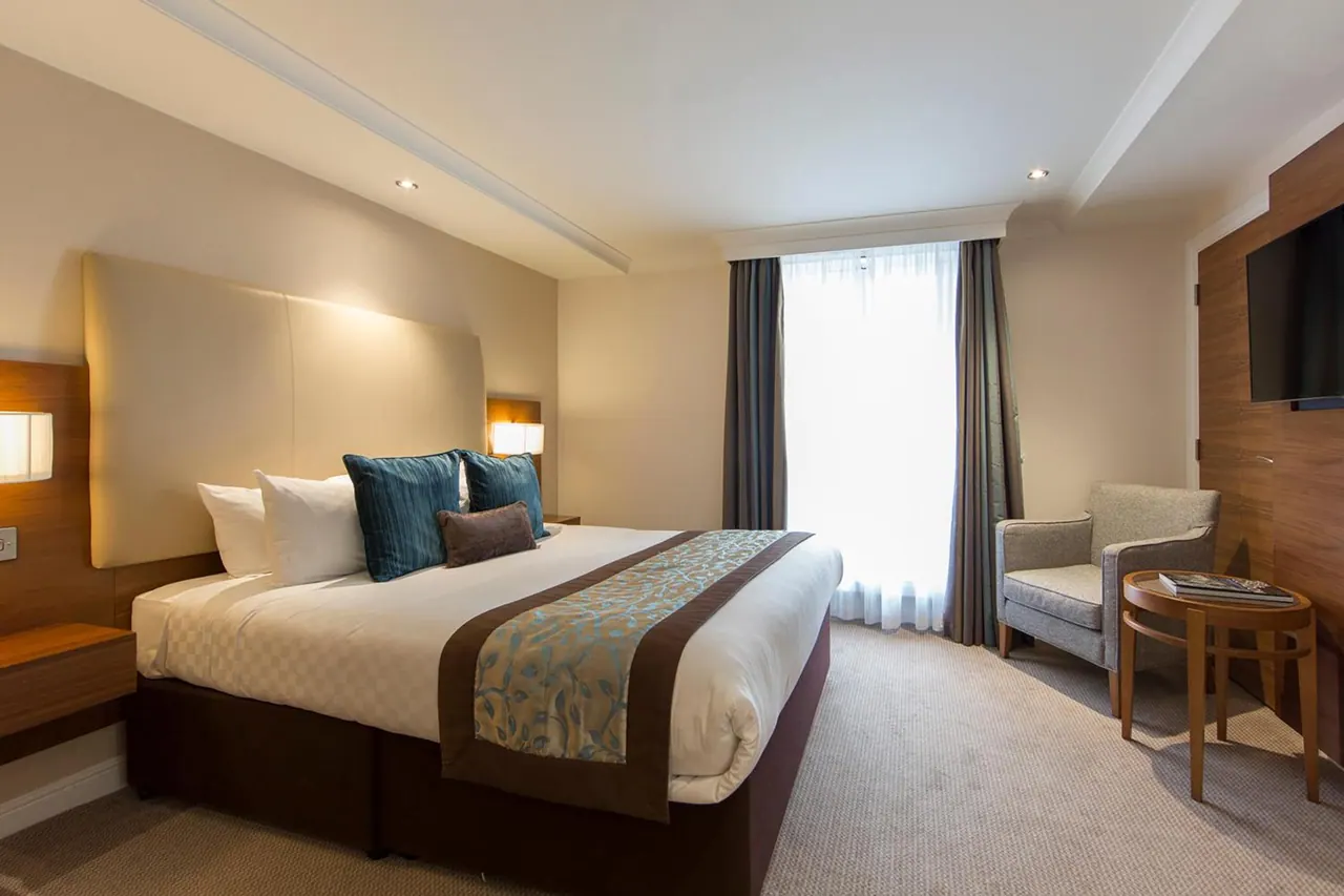 A bedroom with a king bed with teal and white bedding, a grey armchair beside a round wooden side table, and a wall-mounted television.