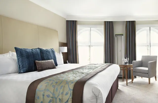 A bedroom with a large made-up bed, blue cushions, arched windows with curtains, and a grey armchair beside a small side table with glasses.