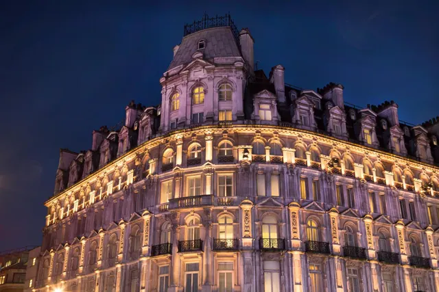 Night view of a multi-storey stone building with a mansard roof, its facade lit from below and windows glowing against a dark blue sky.