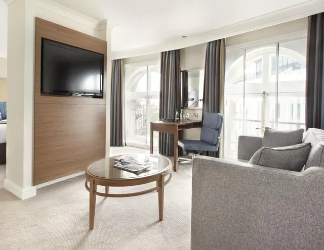 A hotel living area with a wall-mounted television, a grey sofa, a round glass-topped coffee table, and a desk with a chair in front of arched windows looking onto neighbouring buildings.