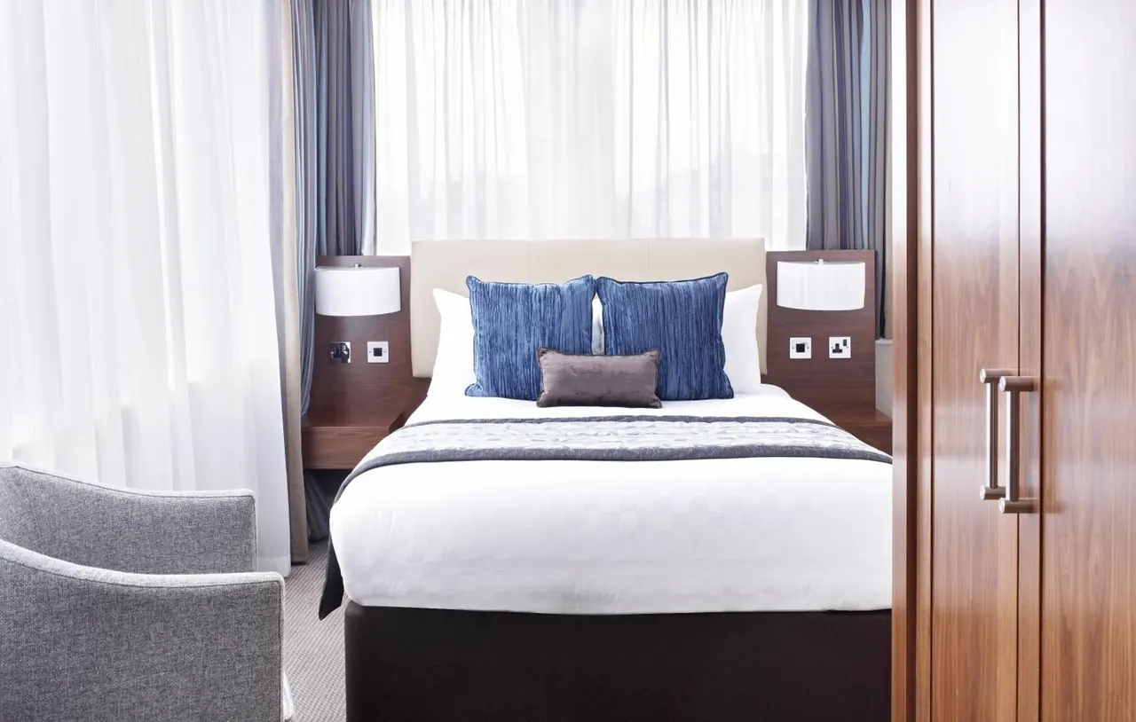 A double bed with blue cushions sits beside a grey armchair in a bedroom with white sheer curtains and a wooden wardrobe.