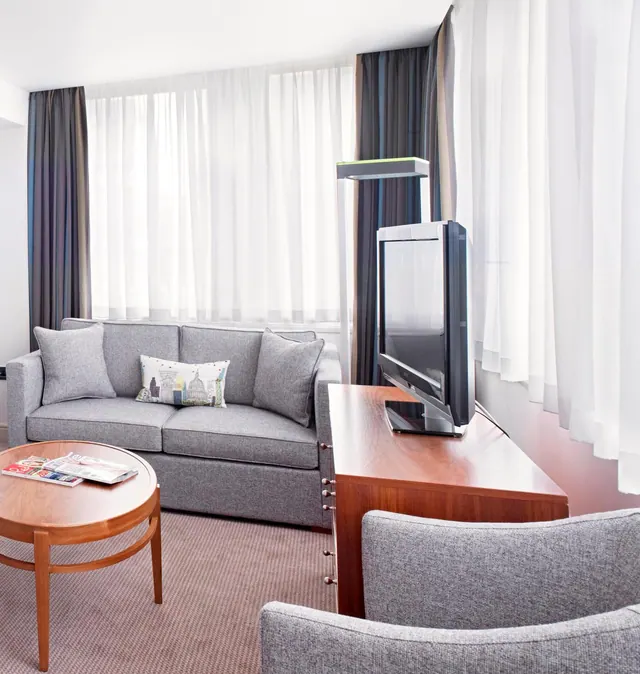 A grey sofa with cushions, a wooden coffee table with magazines, a grey armchair and a flat-screen television on a wooden cabinet sit beside tall sheer white curtains.